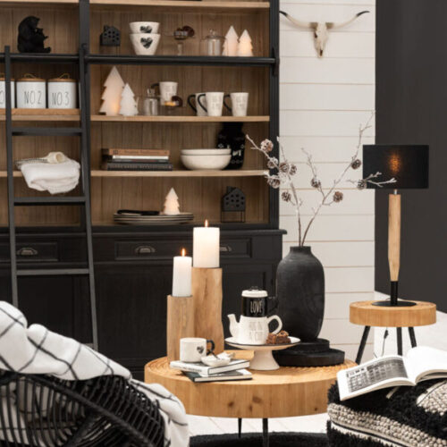 bibliothèque en bois noir et naturel avec échelle en bois noir vu dans un salon cosy table basse en bois naturel avec 2 supports bougies en bois naturel lampe sur pied en bois naturel et chapeau noir, fauteuil en rotin noir et plusieurs bibelots sur étagères de la bibliothèque de couleur blanc et noir