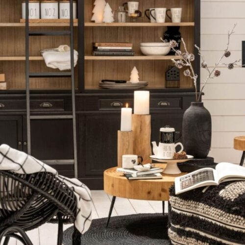bibliothèque en bois noir et naturel avec échelle en bois noir vu dans un salon cosy table basse en bois naturel avec 2 supports bougies en bois naturel lampe sur pied en bois naturel et chapeau noir, fauteuil en rotin noir et plusieurs bibelots sur étagères de la bibliothèque de couleur blanc et noir