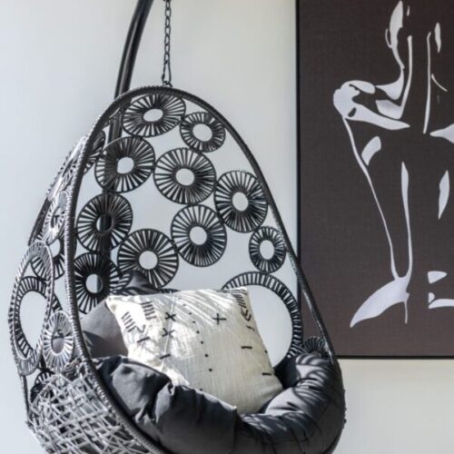 fauteuil suspendu en rotin noir avec des cercles noirs contenant des coussins noirs devant un cadre noir représentant une femme assise sur un mur blanc