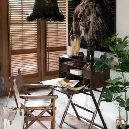 secrétaire bureau en bois de manguier marron ouvert avec un grand cadre en verre trempé représentant une tête de lion sur fond noir un fauteuil en cuir et bois de manguier est installé devant le bureau une suspension en herbes et roseau de couleur noire est situé au dessus du coin bureau
