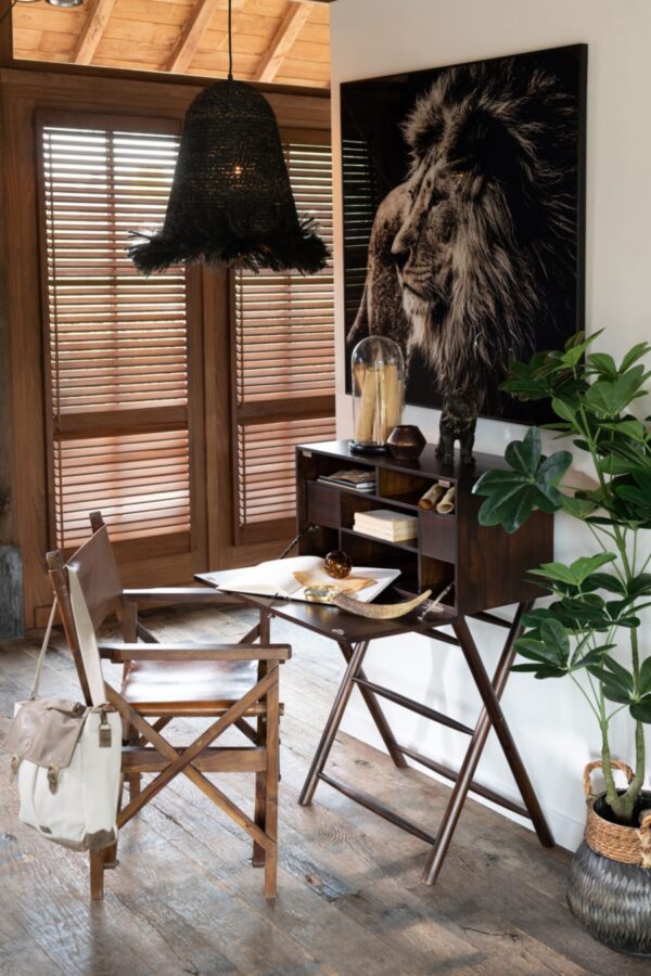 secrétaire bureau en bois de manguier marron ouvert avec un grand cadre en verre trempé représentant une tête de lion sur fond noir un fauteuil en cuir et bois de manguier est installé devant le bureau une suspension en herbes et roseau de couleur noire est situé au dessus du coin bureau