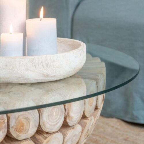 table basse ronde en teck naturel avec plateau en verre sur un tapis beige avec des bougies blanches posées dessus