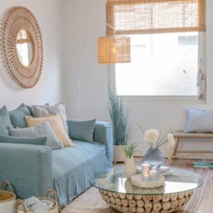 table basse ronde avec des disques de teck et un plateau en verre dans un salon dans les couleurs bleu beige et naturel avec un canapé bleu et un miroir rond en rotin et une suspension en bambou