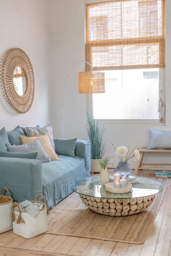 table basse ronde avec des disques de teck et un plateau en verre dans un salon dans les couleurs bleu beige et naturel avec un canapé bleu et un miroir rond en rotin et une suspension en bambou