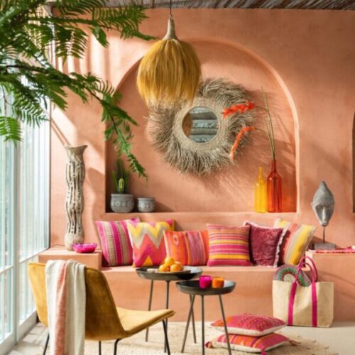 suspension en herbes naturelles miroir rond en roseau et herbes naturelles statue figurine ethnique sur socle vase haut en bois fauteuil lounge en daim ocre et pieds en métal tables gigognes en métal noir coussins colorés dans les teintes roses orangées sac en tissu rose et beige vase bouteille orange translucide avec arbre artificiel