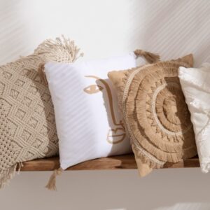 étagère murale en bois d'acacia avec 4 coussins posés dessus un coussin macramé en coton beige avec losanges en relief un coussin visage beige en polyester blanc un coussin en polyester beige avec soleil de tissus en relief un coussin blanc avec croix en relief