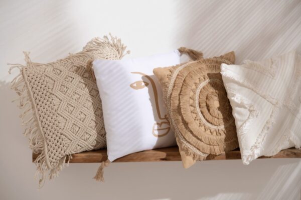 étagère murale en bois d'acacia avec 4 coussins posés dessus un coussin macramé en coton beige avec losanges en relief un coussin visage beige en polyester blanc un coussin en polyester beige avec soleil de tissus en relief un coussin blanc avec croix en relief