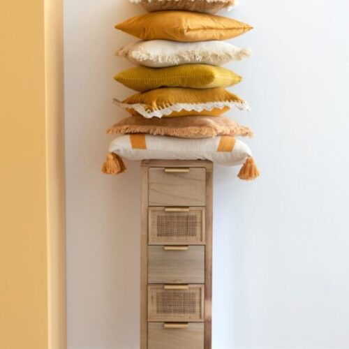 petite armoire fine en bois et rotin naturel tas de coussins posés dessus dans les teintes ocre orangées