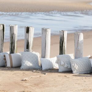 coussin macramé rectangulaire en coton beige avec losanges sur le milieu en relief sur une plage au bord de l'eau posé sur le sable aligné avec d'autres coussins
