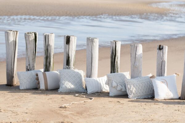 coussin macramé rectangulaire en coton beige avec losanges sur le milieu en relief sur une plage au bord de l'eau posé sur le sable aligné avec d'autres coussins