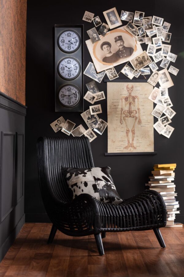 fauteuil lounge en rotin noir vu de biais avec un coussin en peau de vache posé dessus dans un salon mur noir et sol plancher marron beaucoup de photos anciennes au mur et une horloge vintage